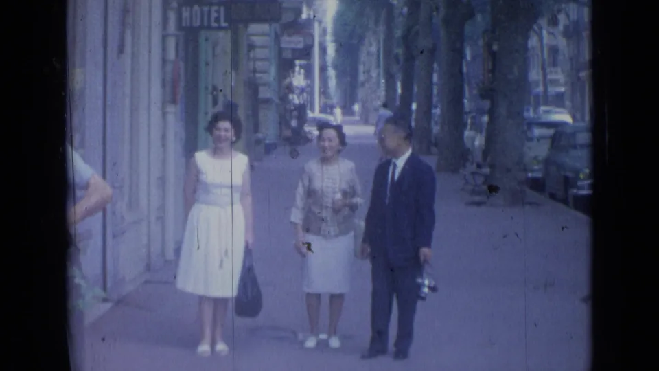 Three adults in 1960s clothing stand or slow on a Cannes sidewalk, framed by storefronts, trees, and parked cars.