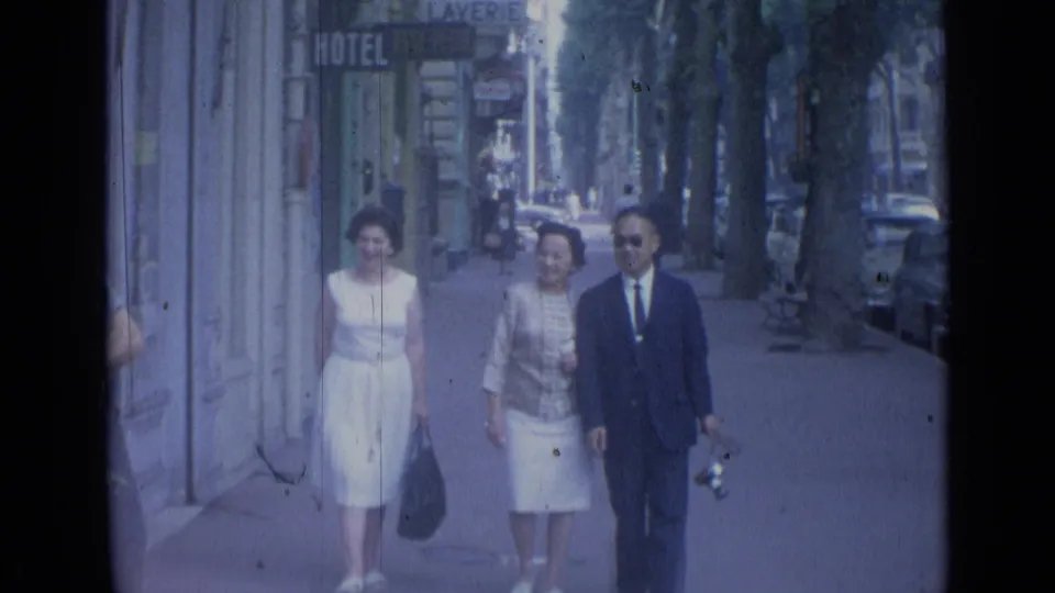 Three adults smile and walk together on a Cannes sidewalk, with shop signs, trees, and parked cars visible in the 8mm frame.