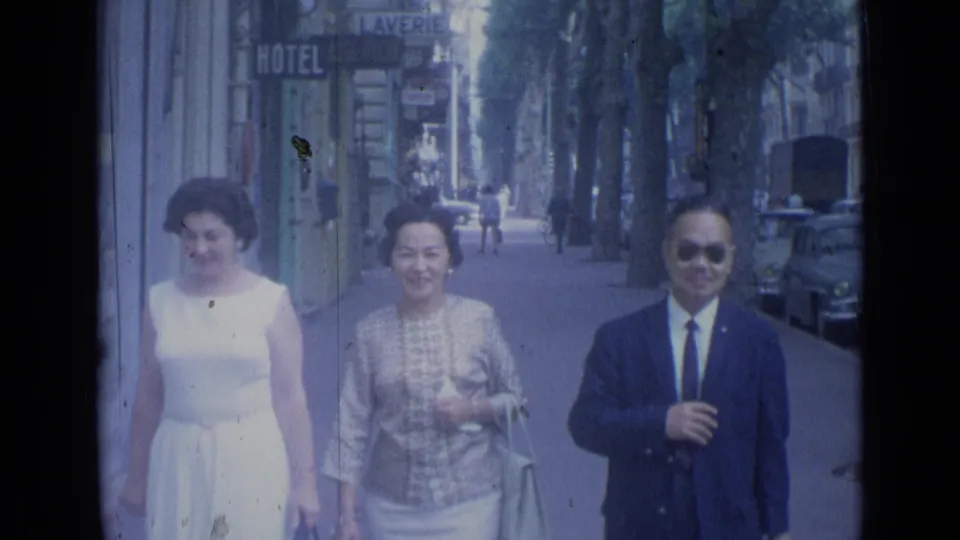 In the 1962 Cannes home-movie scene, the adults smile while approaching, with hotel signage and parked cars behind them.