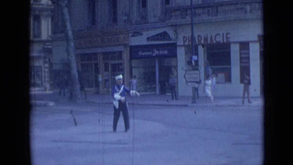 A uniformed traffic officer directs or monitors a Cannes street in 1962, with pharmacy lettering, pedestrians, and stone-front buildings behind him.