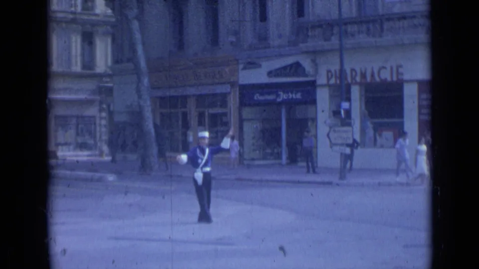 A Cannes street view from 1962 shows a uniformed traffic officer in the roadway, pedestrians by shopfronts, and visible pharmacy signage.
