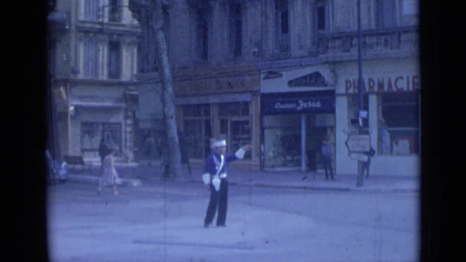 1962 Cannes traffic officer directs vehicles in front of pharmacy and shop signs, with pedestrians crossing the background.
