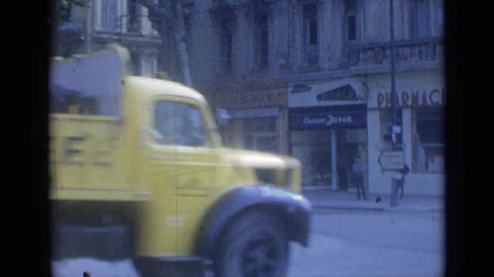 A yellow commercial truck crosses the foreground of the 1962 Cannes street scene, obscuring part of the officer and pharmacy storefront.