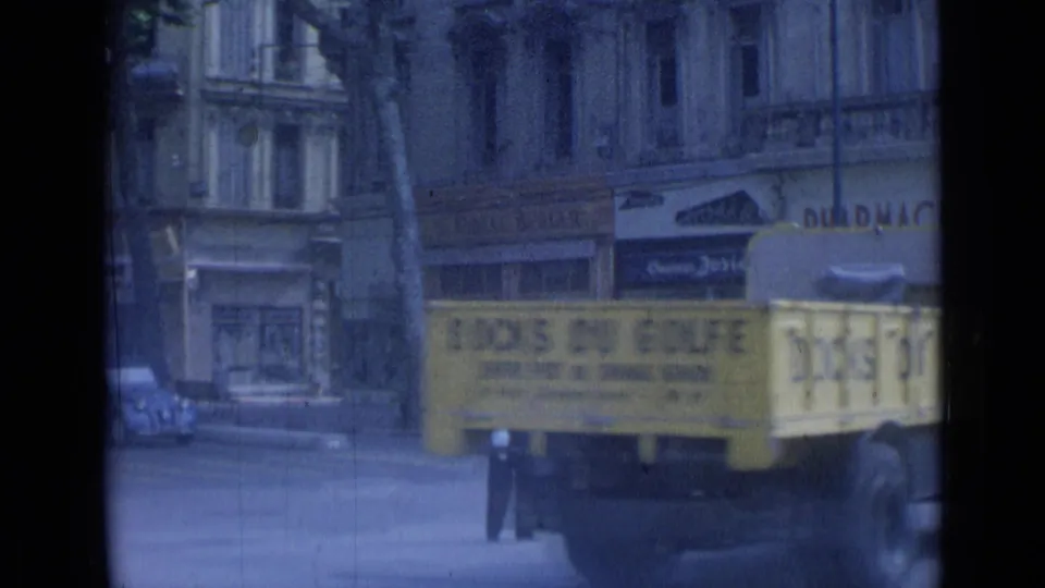The yellow truck continues through the 1962 Cannes street frame, with the traffic officer partly hidden and pharmacy signage behind it.