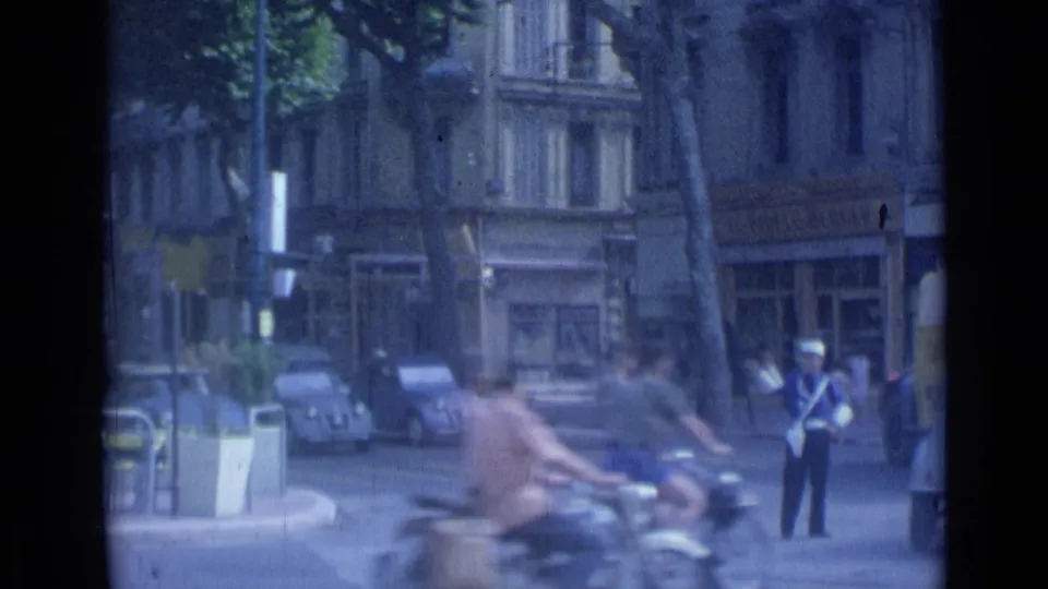 Cannes traffic scene with blurred scooters in the foreground, parked cars, storefronts, and a uniformed traffic policeman.