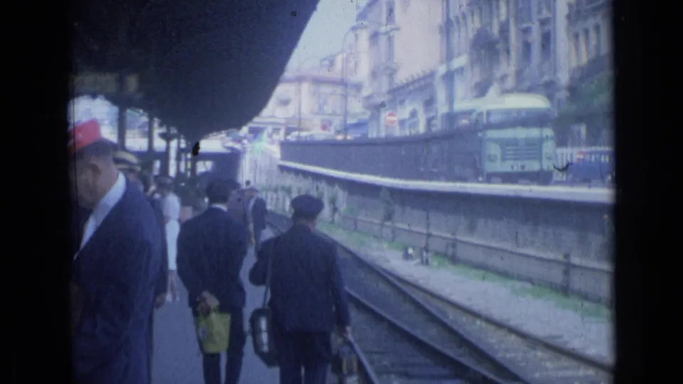 1962 Cannes railway-side street view with pedestrians, tracks, a long platform wall, and urban buildings beyond.