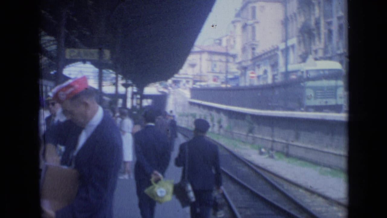 Crowded station platform beside tracks as a man in a red cap stands near waiting passengers.