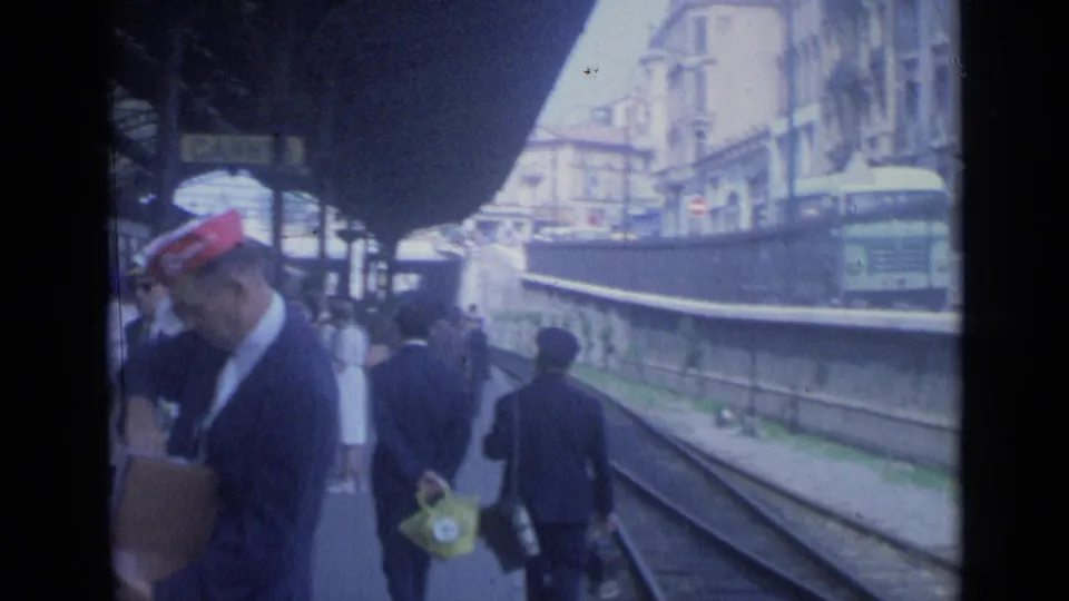 1962 Cannes station platform with travelers waiting near tracks while a red-capped railway worker stands in the foreground.