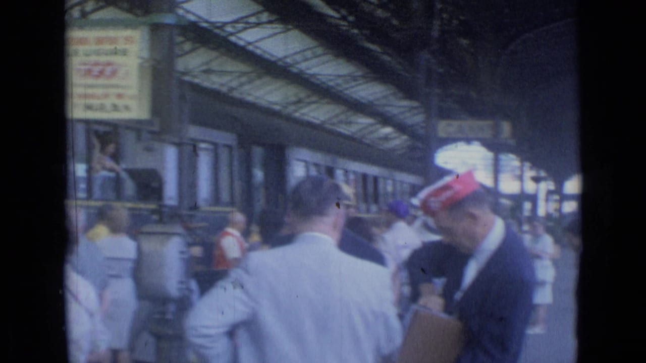 Crowded Cannes rail platform in 1962 with red-capped staff, passengers, overhead signage, and a train at the station.