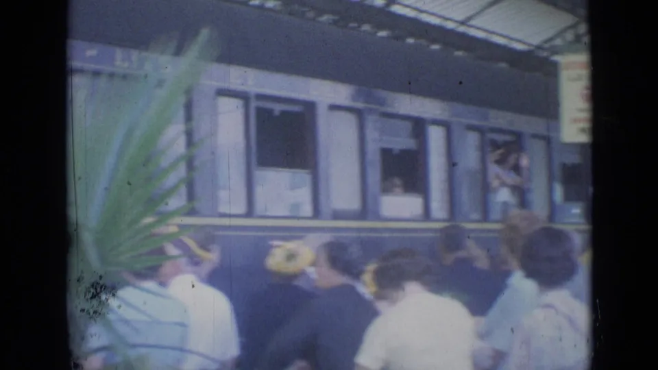 Travelers in 1960s clothing watch a blue passenger railcar from the Cannes station platform.