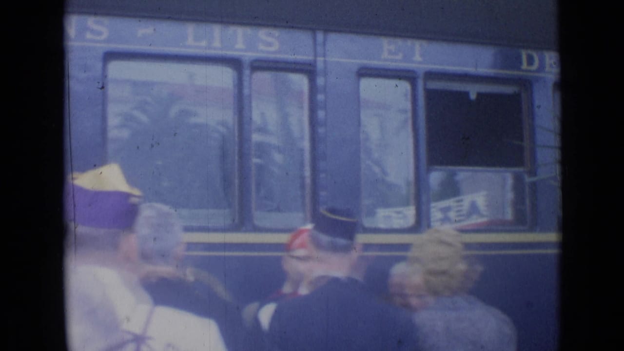 Blue passenger train passing platform crowd with uniform caps in foreground.