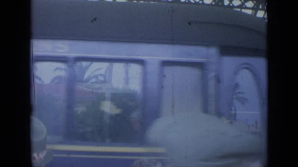 Soft, handheld view of a blue train at the station, with blurred people in the foreground and carriage windows behind them.