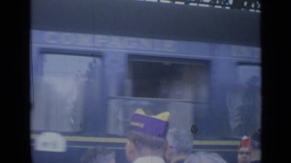Cannes, France 1962 frame with a blue passenger train moving past platform onlookers, including men in distinctive caps.