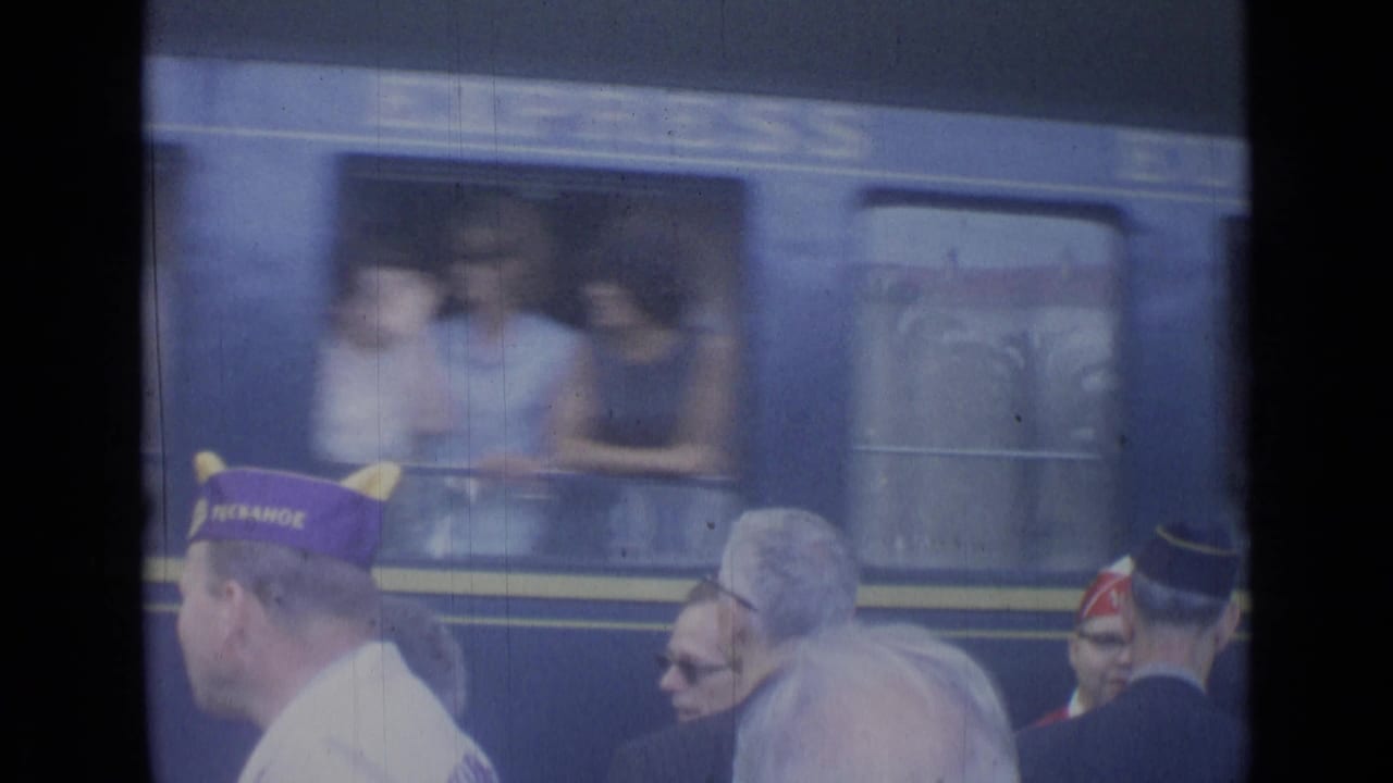 Men in veterans caps stand on a railway platform beside a blue passenger train.