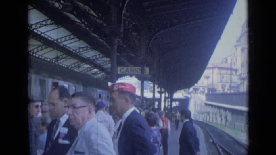 1962 Cannes station platform under the canopy, with suited men, veterans caps, rail tracks, and a visible Cannes sign.