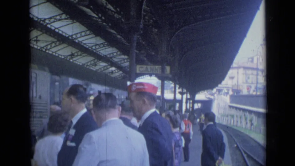 Cannes station platform in 1962 with men in suits and caps gathered beside the tracks under the covered platform.