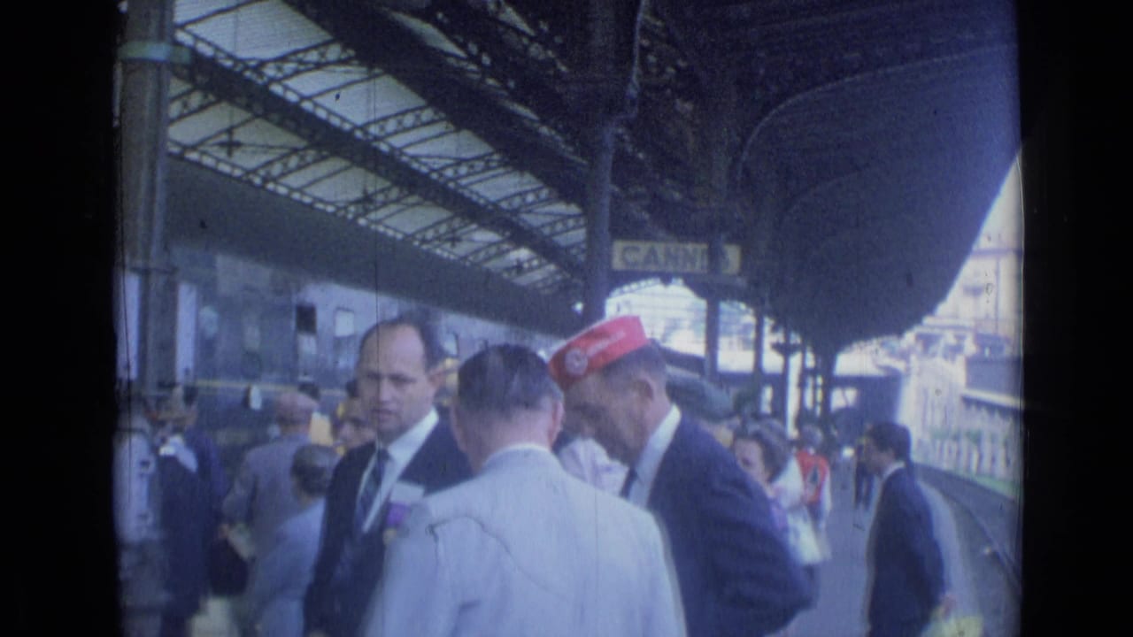 1962 Cannes station platform with suited men, a red-capped worker, passengers, and a train under the canopy.