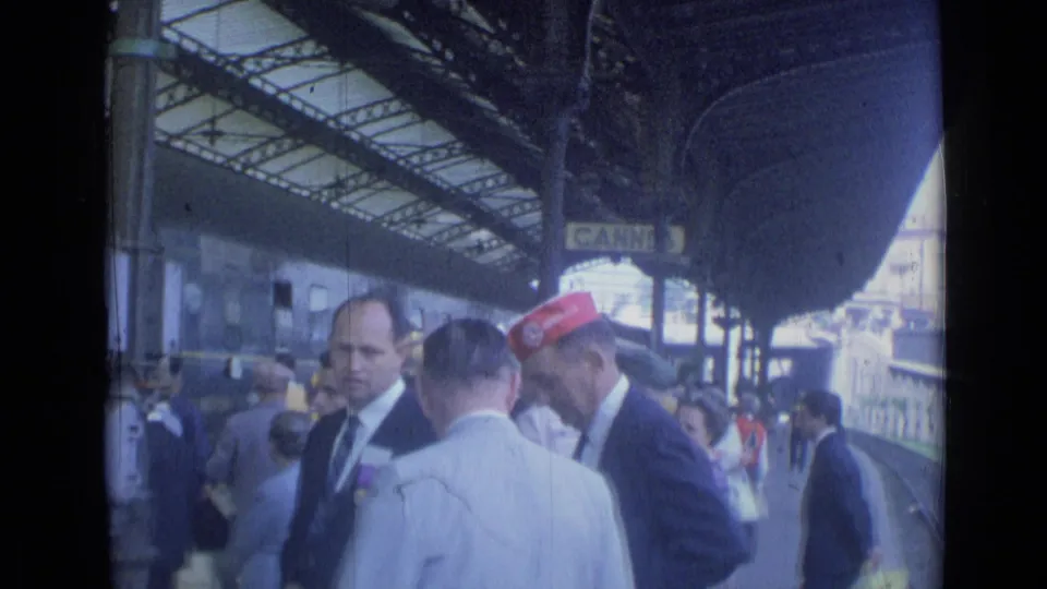 1962 Cannes station platform with suited men, a red-capped worker, passengers, and a train under the canopy.