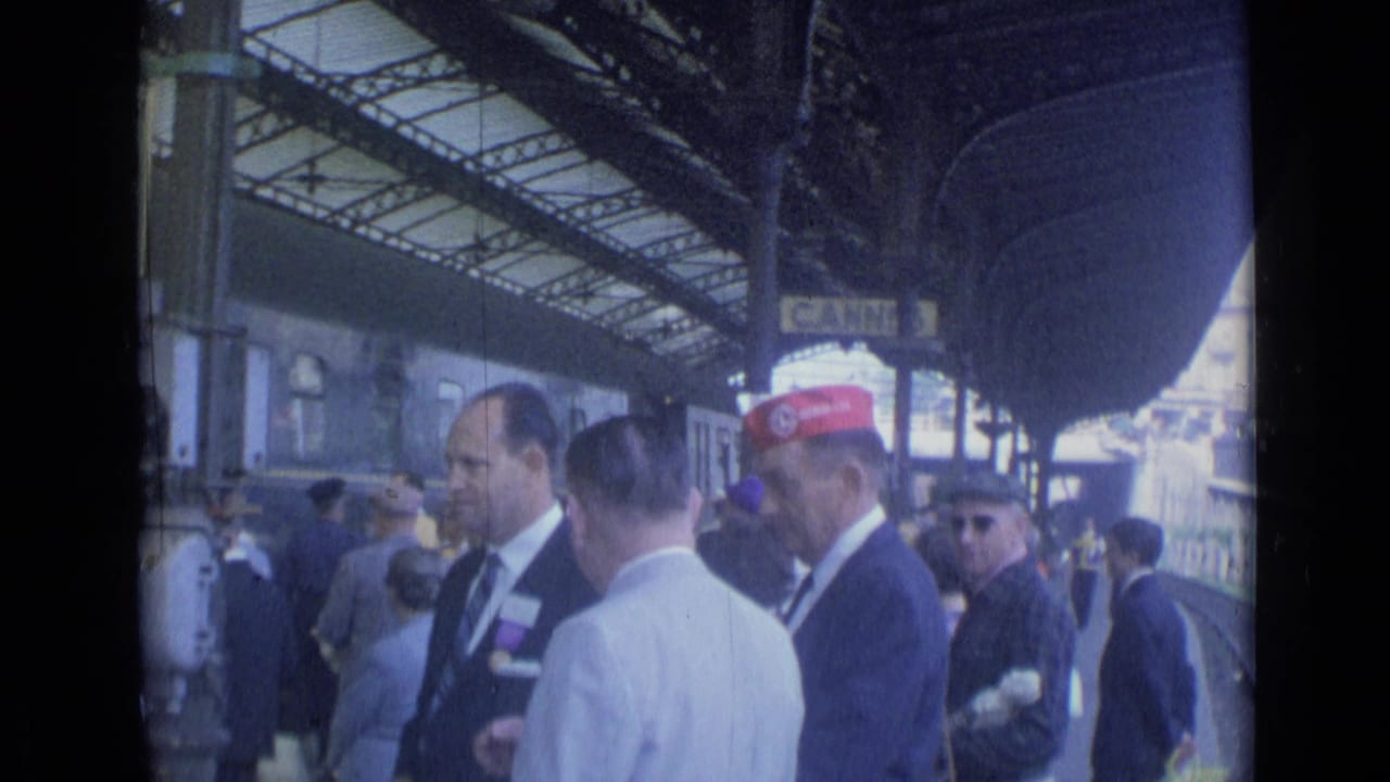 Station platform crowd with suited men, a red-capped worker, and train cars.