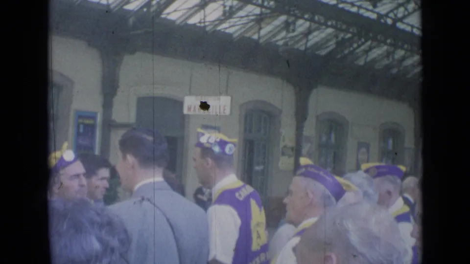 Cannes station platform with a Marseille sign, suited men, and a group wearing matching purple and yellow caps and vests.