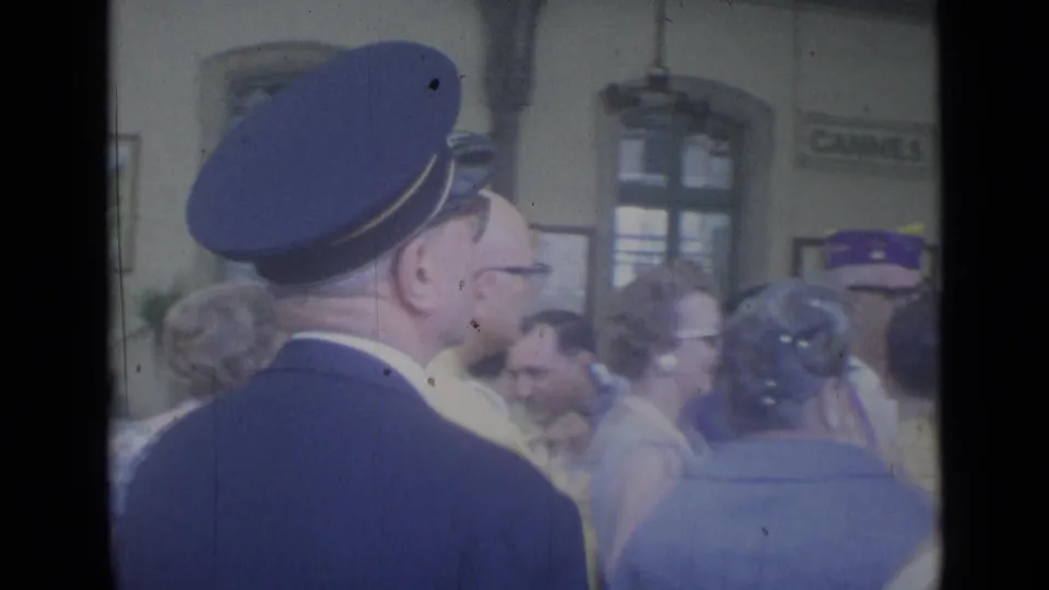 A uniformed station official stands among passengers in the 1962 Cannes railway station concourse.
