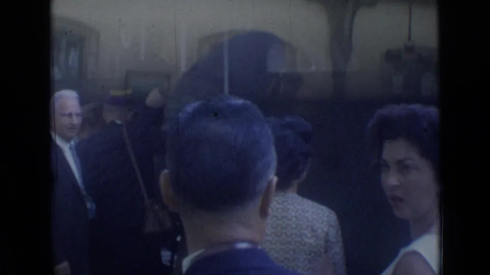 1962 Cannes station crowd seen from a handheld 8mm viewpoint, with suited adults and a woman in a white dress among passengers.