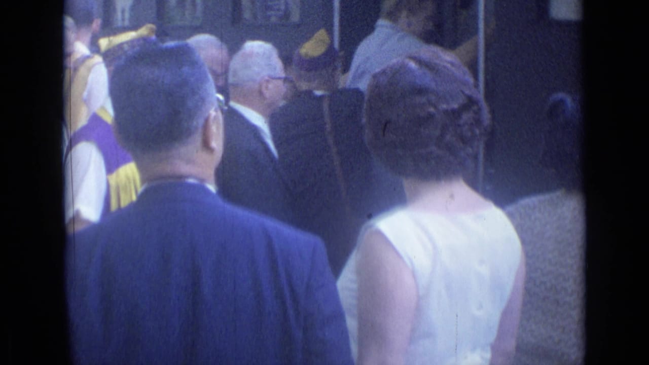 Adults in formal 1960s attire move through a Cannes station platform, with uniformed staff visible near the crowd.