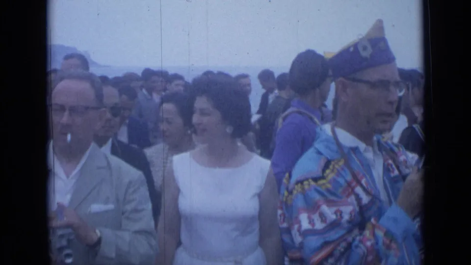 Cannes, France, 1962 crowd scene showing spectators in formal clothes near a costumed participant, with water or hillside faintly behind.