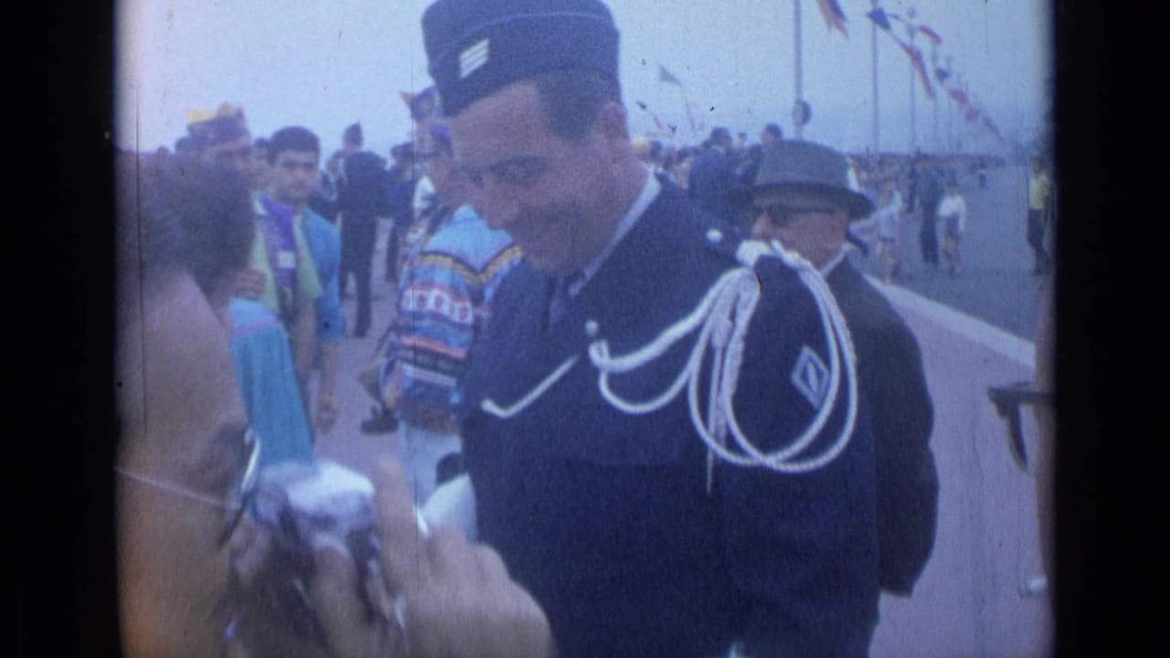 Uniformed man looks down near a camera and surrounding promenade crowd.