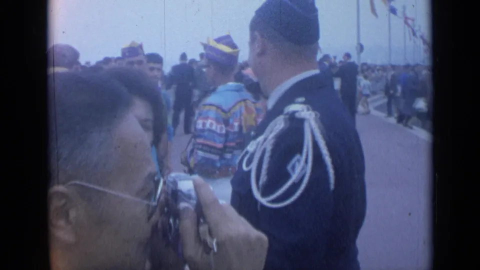 A foreground photographer and a uniformed man seen from behind appear among Cannes promenade spectators in 1962.