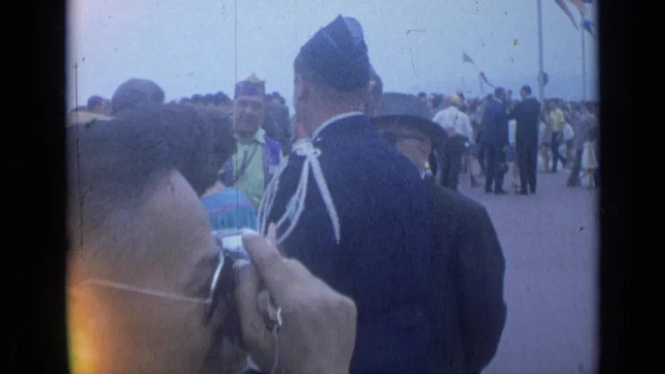 The 1962 Cannes promenade crowd surrounds a uniformed man viewed from behind as a foreground visitor photographs nearby.