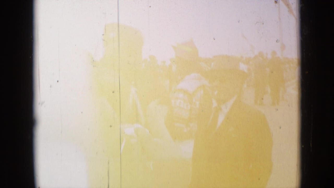 1962 Cannes home-movie frame with a dark-suited man near parade spectators, partly obscured by yellow fading and scratches.