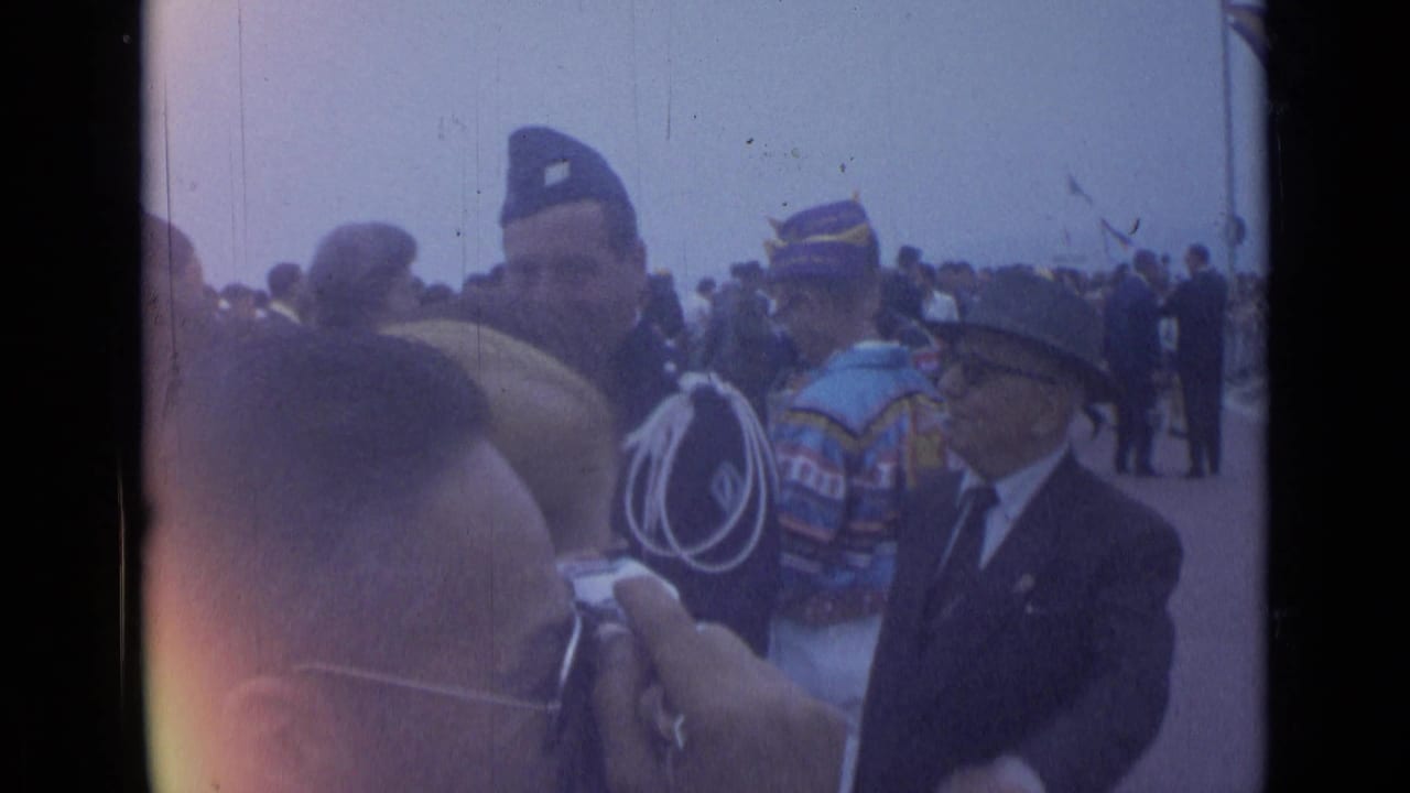 Uniformed men and onlookers gather near a parade route, with a camera hand in front.