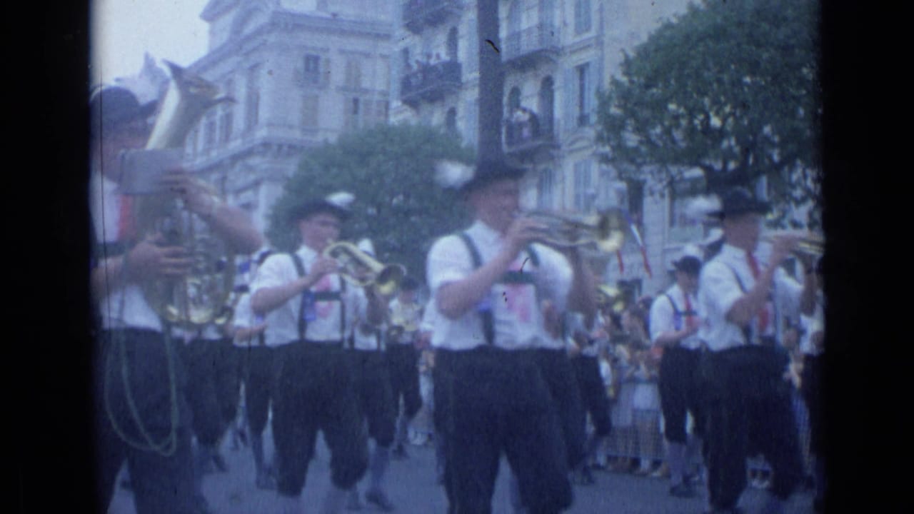 In Cannes in 1962, uniformed brass musicians march past buildings and spectators during a street parade.