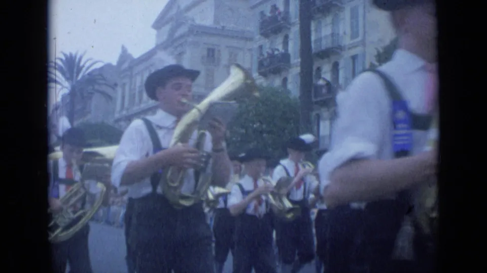 In the Cannes parade, a brass player in a dark hat marches past the camera with fellow musicians and spectators behind.