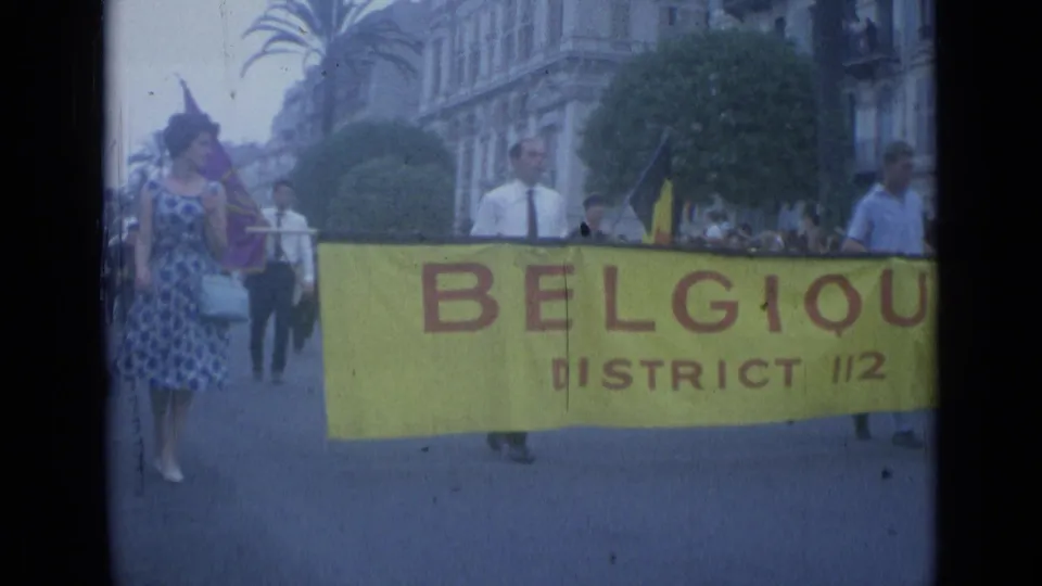 1962 Cannes parade frame with marchers holding a yellow "BELGIQUE DISTRICT 112" banner as spectators watch from the sidewalk.
