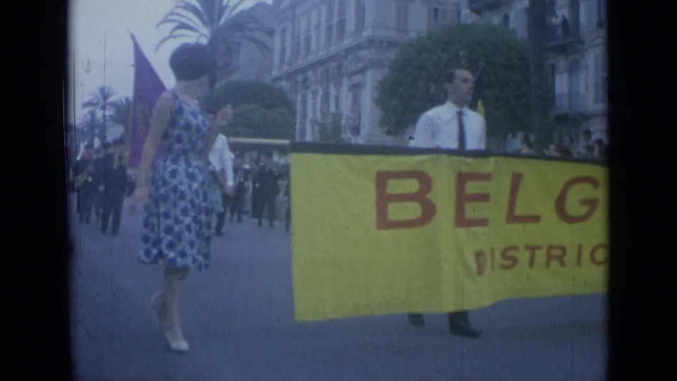 1962 Cannes street parade frame showing a woman in a blue patterned dress walking next to the Belgique banner and flag bearers.