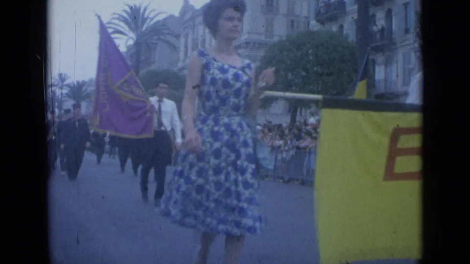 1962 Cannes parade frame with a woman in a blue patterned dress, purple flag, Belgian-colored flag, and yellow banner near the crowd.