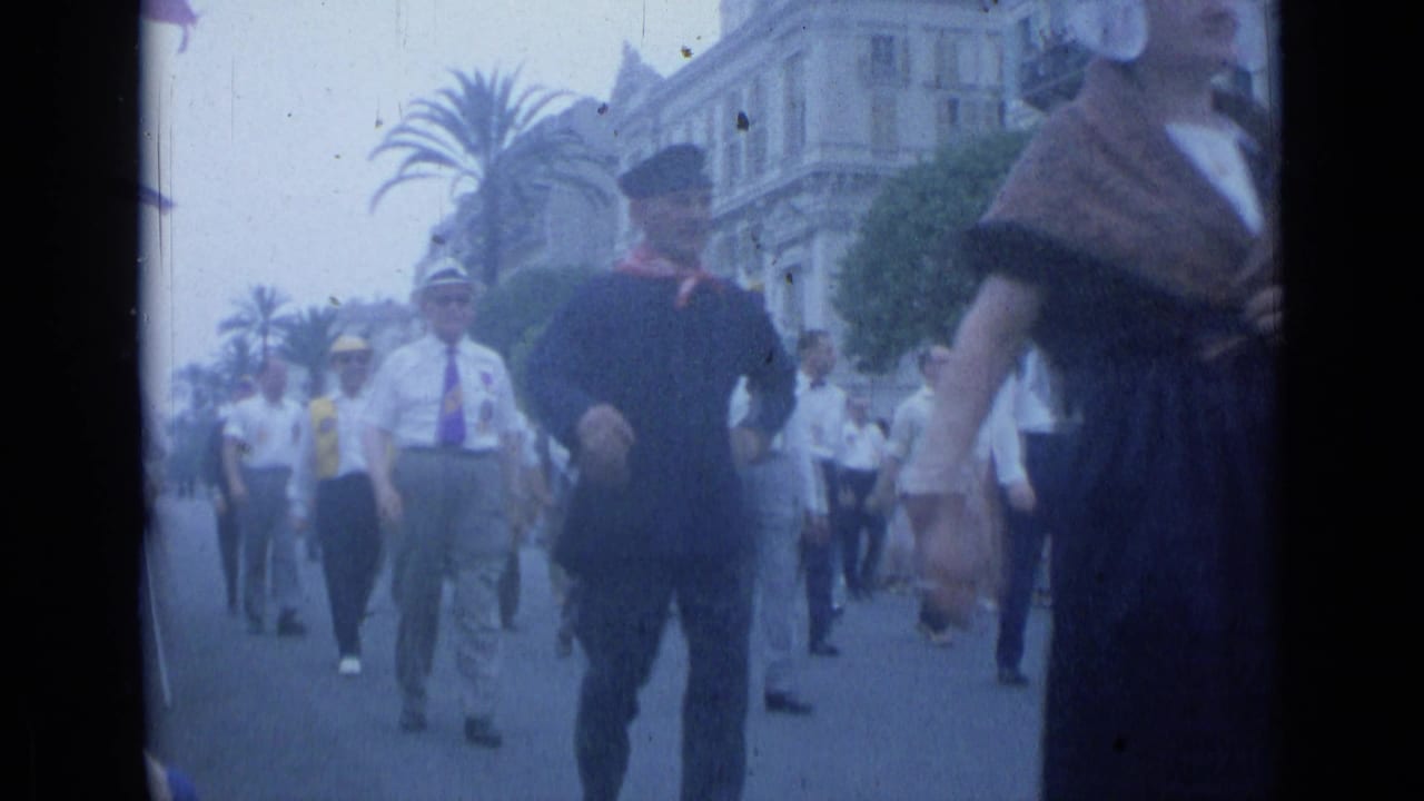 Men in white shirts and caps march on a palm-lined street as a woman in dark clothing passes near the camera.