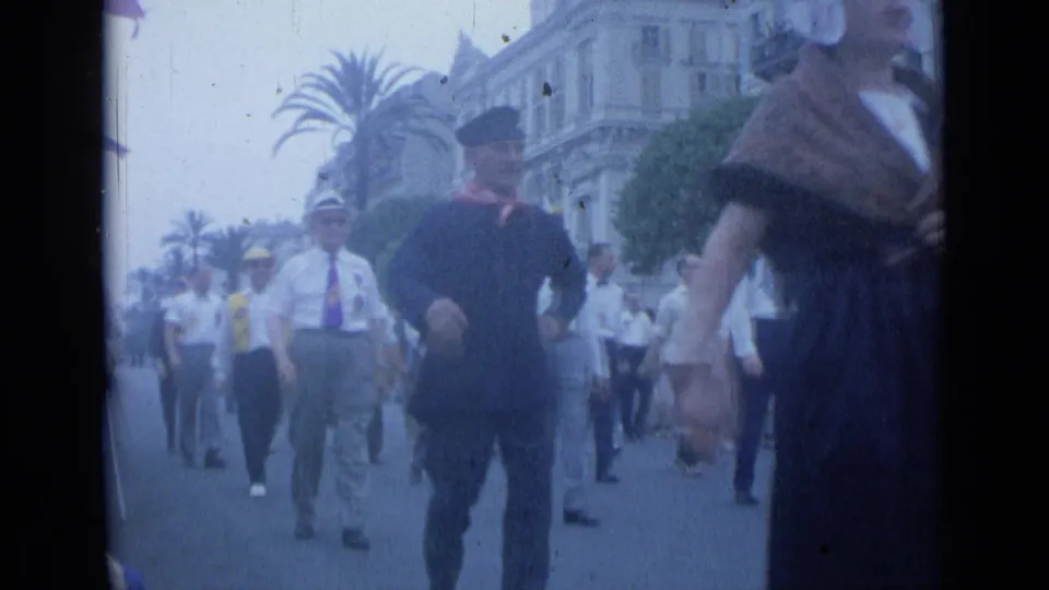 1962 Cannes home-movie frame showing marching men in white shirts and caps, with a woman in dark dress near the foreground.