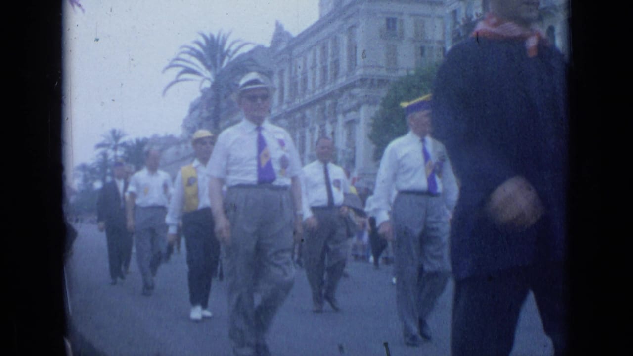 Men in white shirts, ties, and caps walk in formation along a broad street with palm trees and buildings behind them.