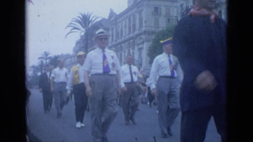 1962 Cannes frame of uniformed-looking marchers in white shirts and caps moving past palm trees and European street facades.