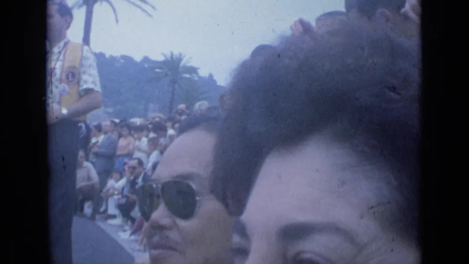 1962 Cannes home-movie frame with a close face in the foreground, a man in sunglasses, and a seated crowd behind them.
