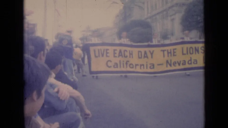 In Cannes, France in 1962, spectators sit near the curb as marchers carry a large California-Nevada Lions banner past buildings.