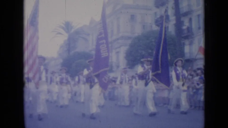 White-uniformed marchers with flags proceed through Cannes during the 1962 parade, with spectators and palm-lined buildings nearby.