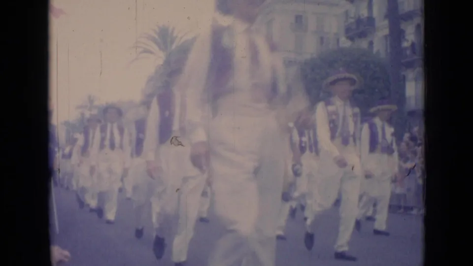 1962 Cannes home-movie frame showing costumed men in white trousers, dark vests and hats parading past palm trees and city buildings.