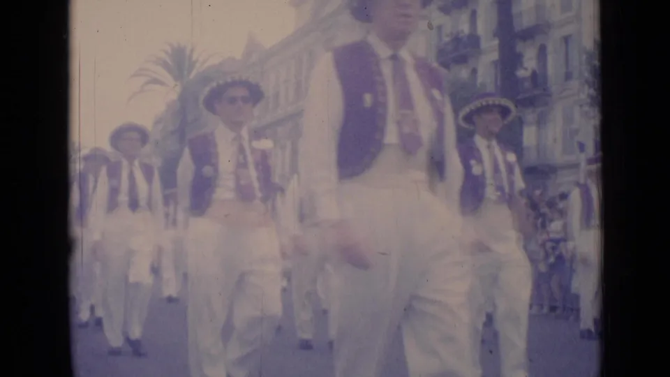 1962 Cannes parade footage with men in coordinated white clothing and hats moving toward the handheld camera on an urban street.