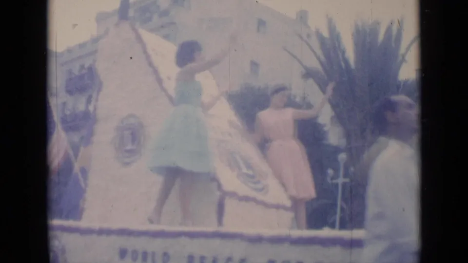 Women in pastel dresses appear beside a Cannes parade float bearing a Lions Club emblem and large plant decoration.