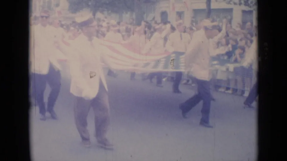 Men in light-colored suits carry a large American flag along a Cannes parade route with spectators behind them.