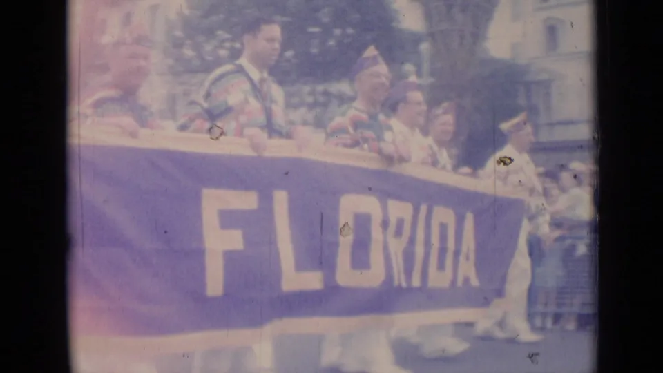 1962 Cannes parade footage shows striped-costume participants carrying a large purple Florida banner past spectators.